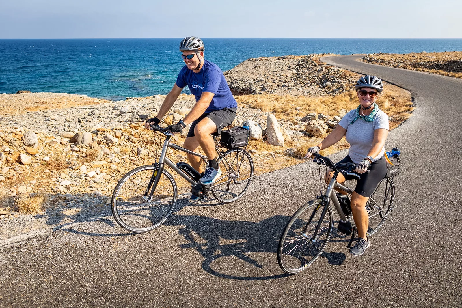 Two guests biking alongside golden coastline, smiling at camera.