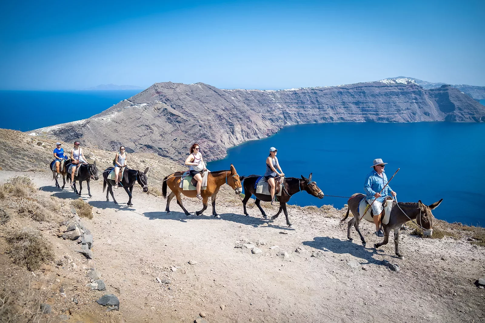 Group of guest on horseback, walking along arid cliffside, ocean below.