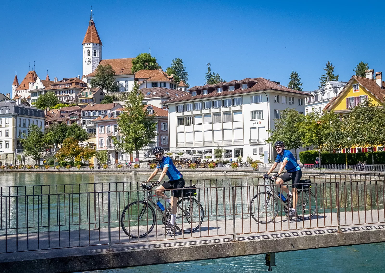 Two guests cycling over concrete bridge, Thun cityscape in distance.
