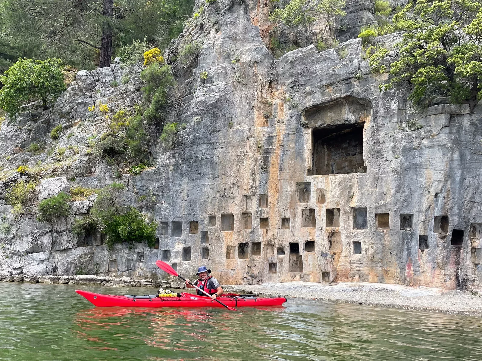 Guest in red kayak, bored out rock wall with square holes behind him.