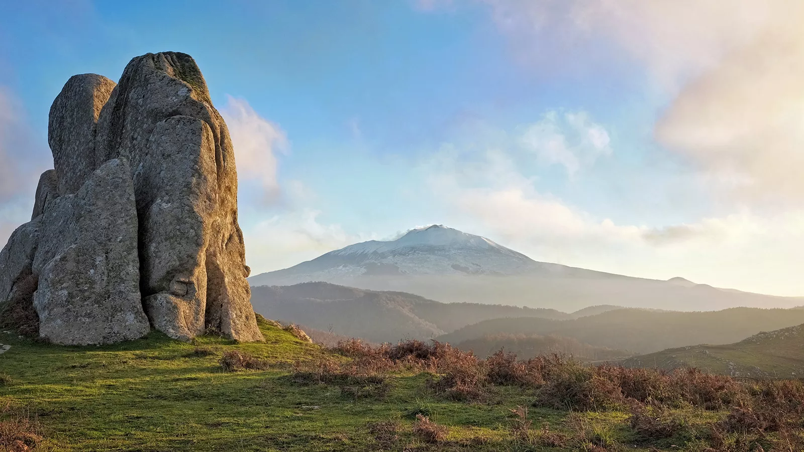 Shot of large rock face, mountains, sunset in distance.