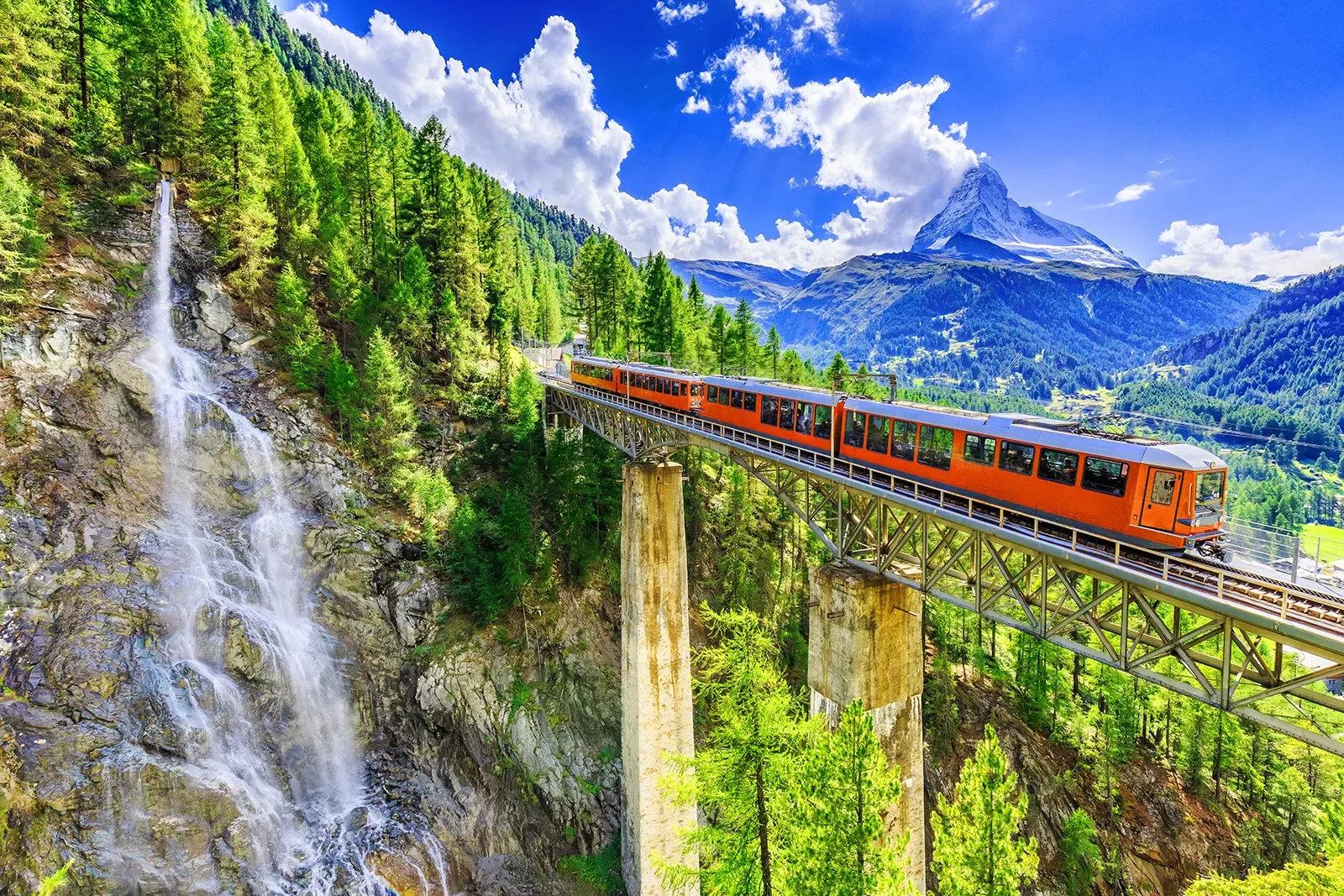 Wide shot of mountain train passing bridge, forest, mountains, sky in distance.