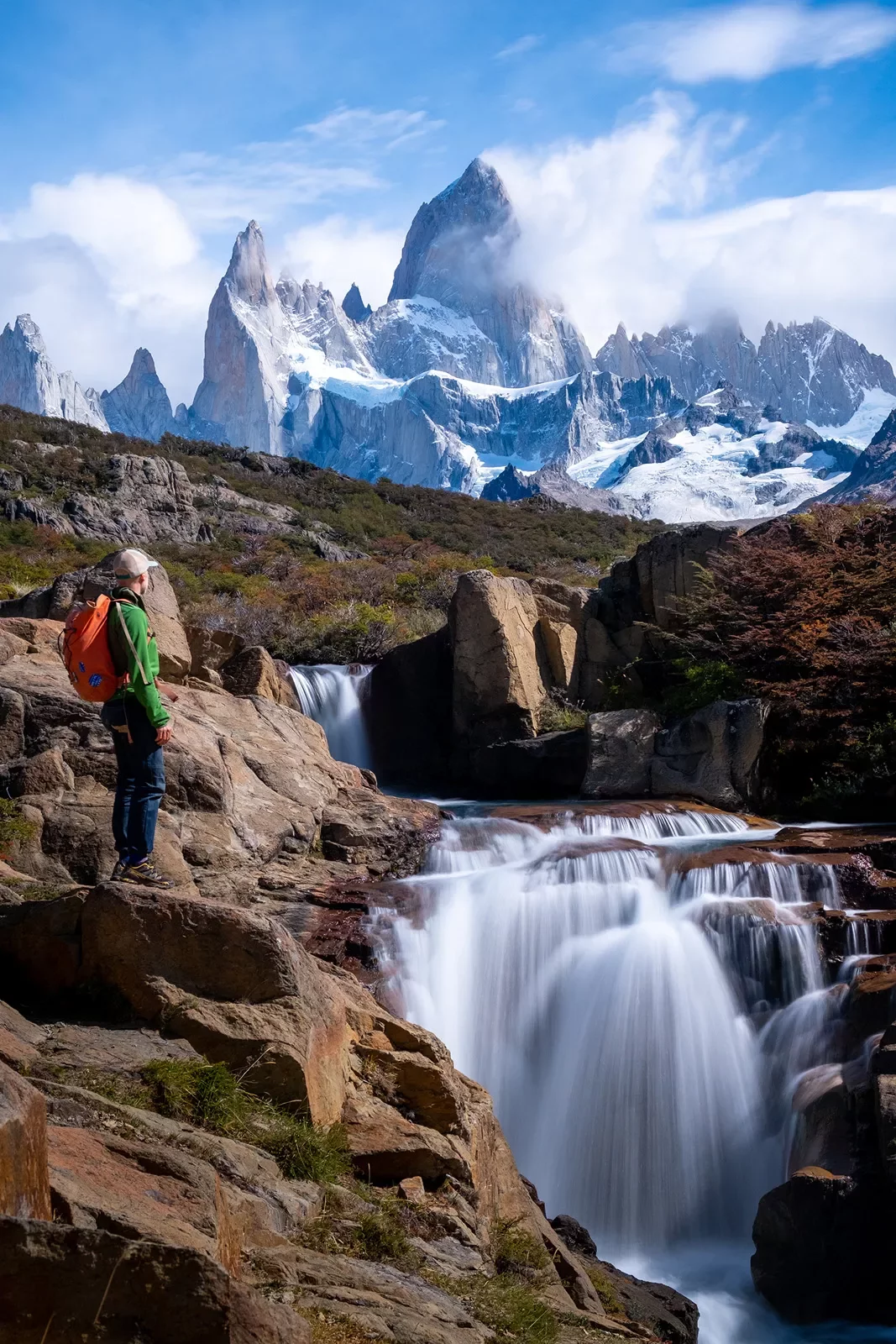 Guest standing next to flowing waterfall, sharp, snowy peaks in background.