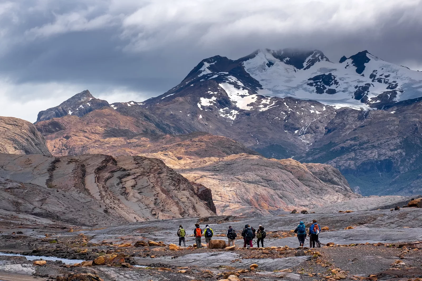 Group of guests walking among arid mountain range, snowy peaks in distance.