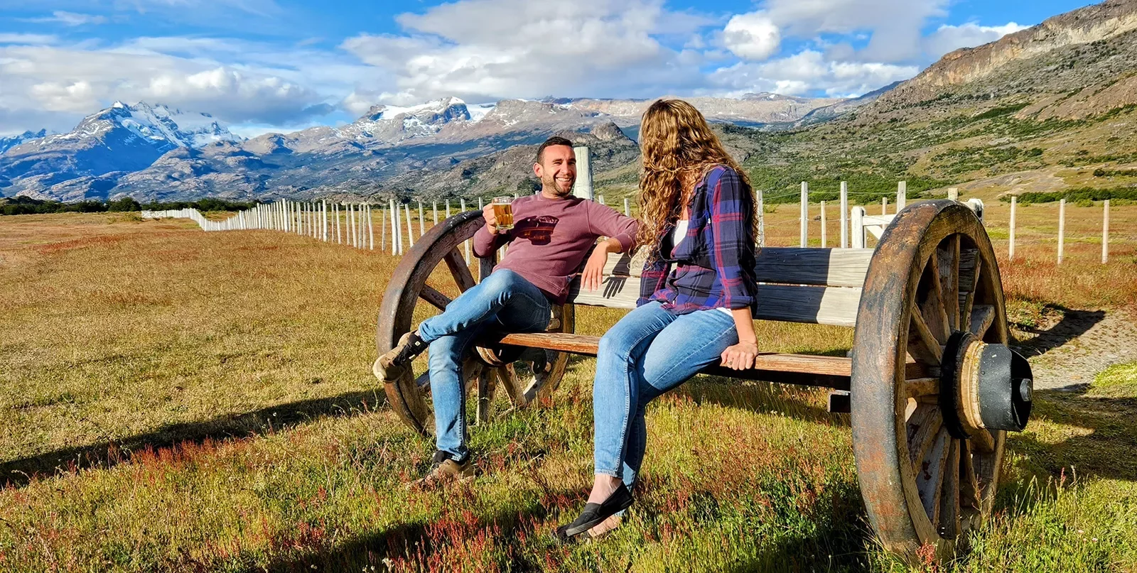 Two guests sitting on wooden bench among golden field, ring of mountains behind them.