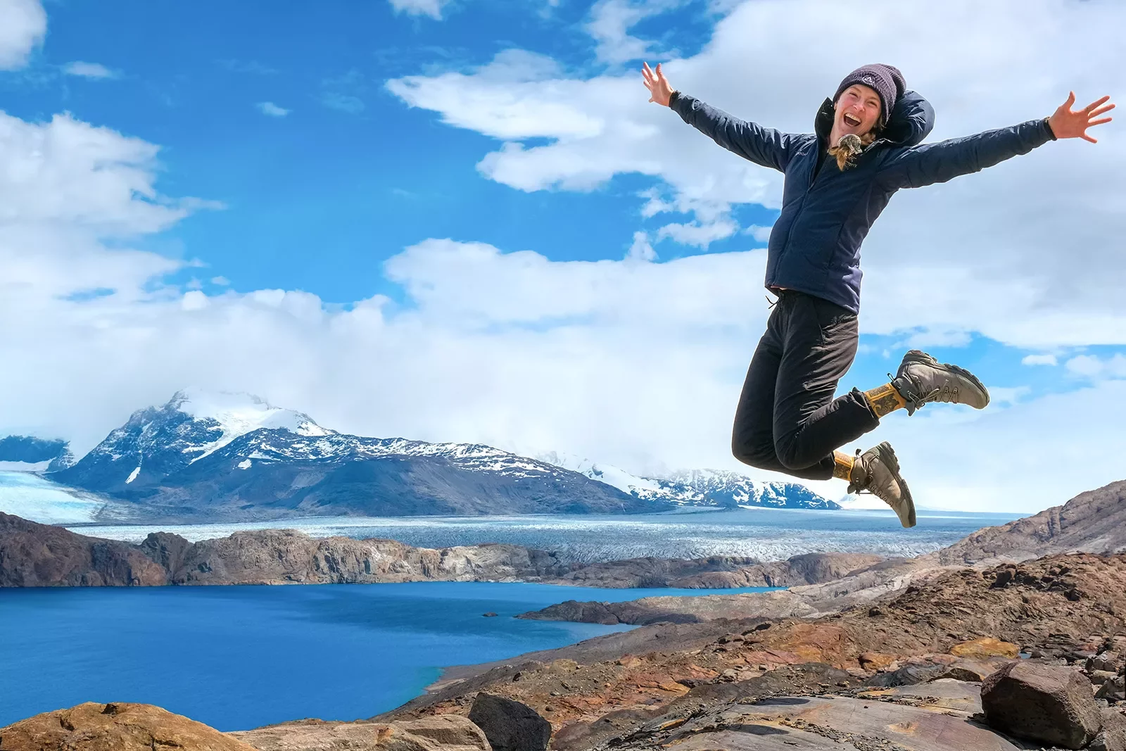 Guest on cliffside, jumping for joy. Lake, snowy mountains behind her.