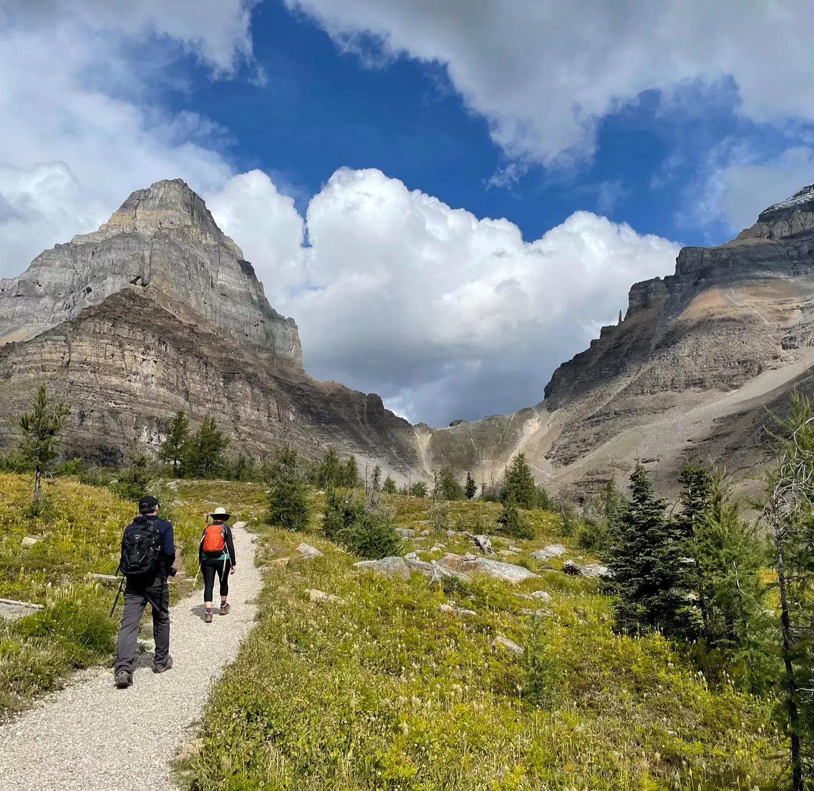 Two guests walking down meadow trail, two large mountain faces in distance.