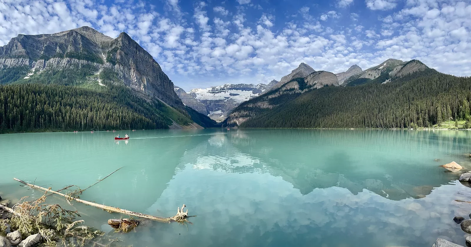 Wide shot of large blue lake, people canoeing in distance, mountains, clouds, sky in background.