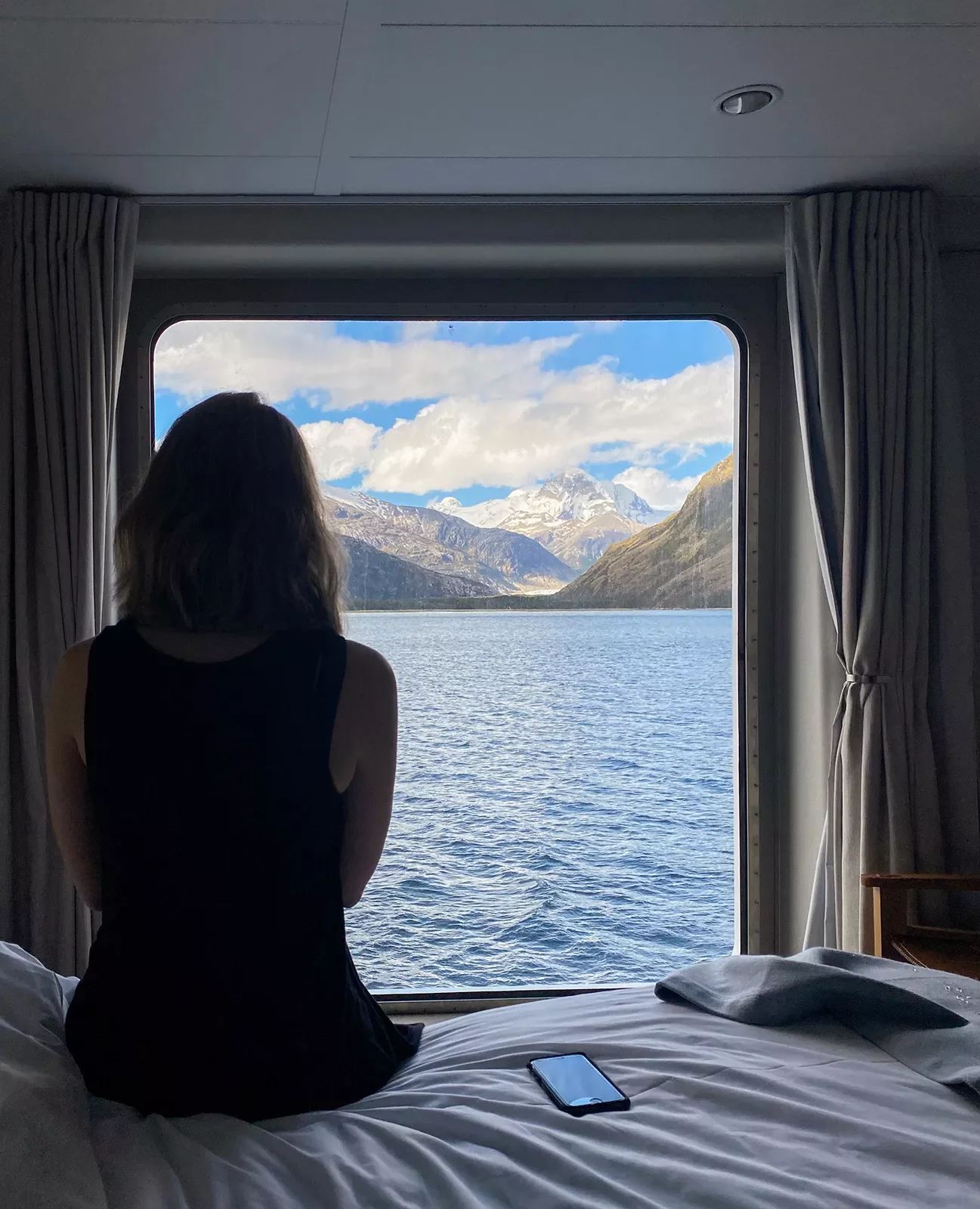 Guest on cruise ship in room, looking out towards mountains.