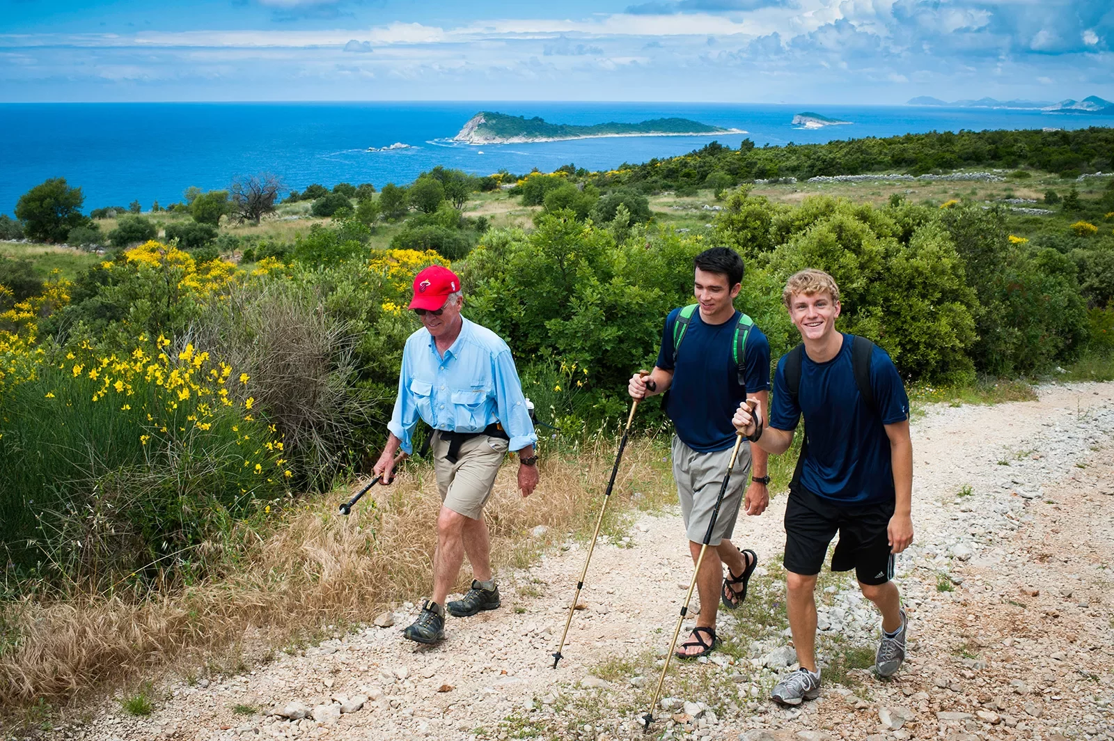 Three guests with hiking poles on coastal trail, ocean, small island chain in distance.