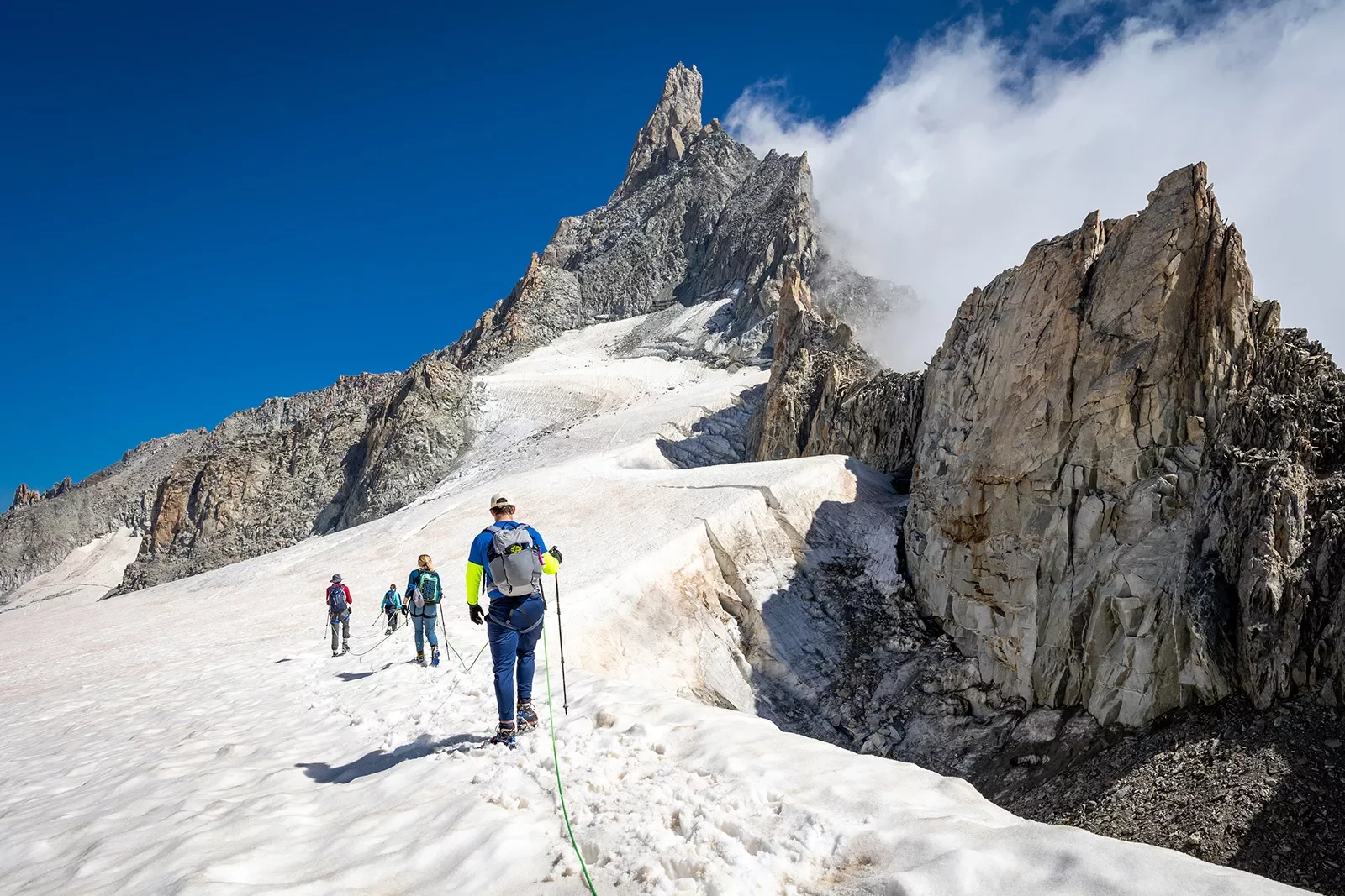 Four guests walking beside snowy cliff, sharp, craggy mountain behind them.