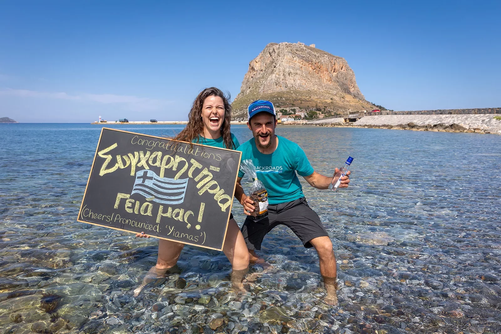 Two leaders holding Greek lettered &quot;Congratulations&quot; signage, standing in shallow water.