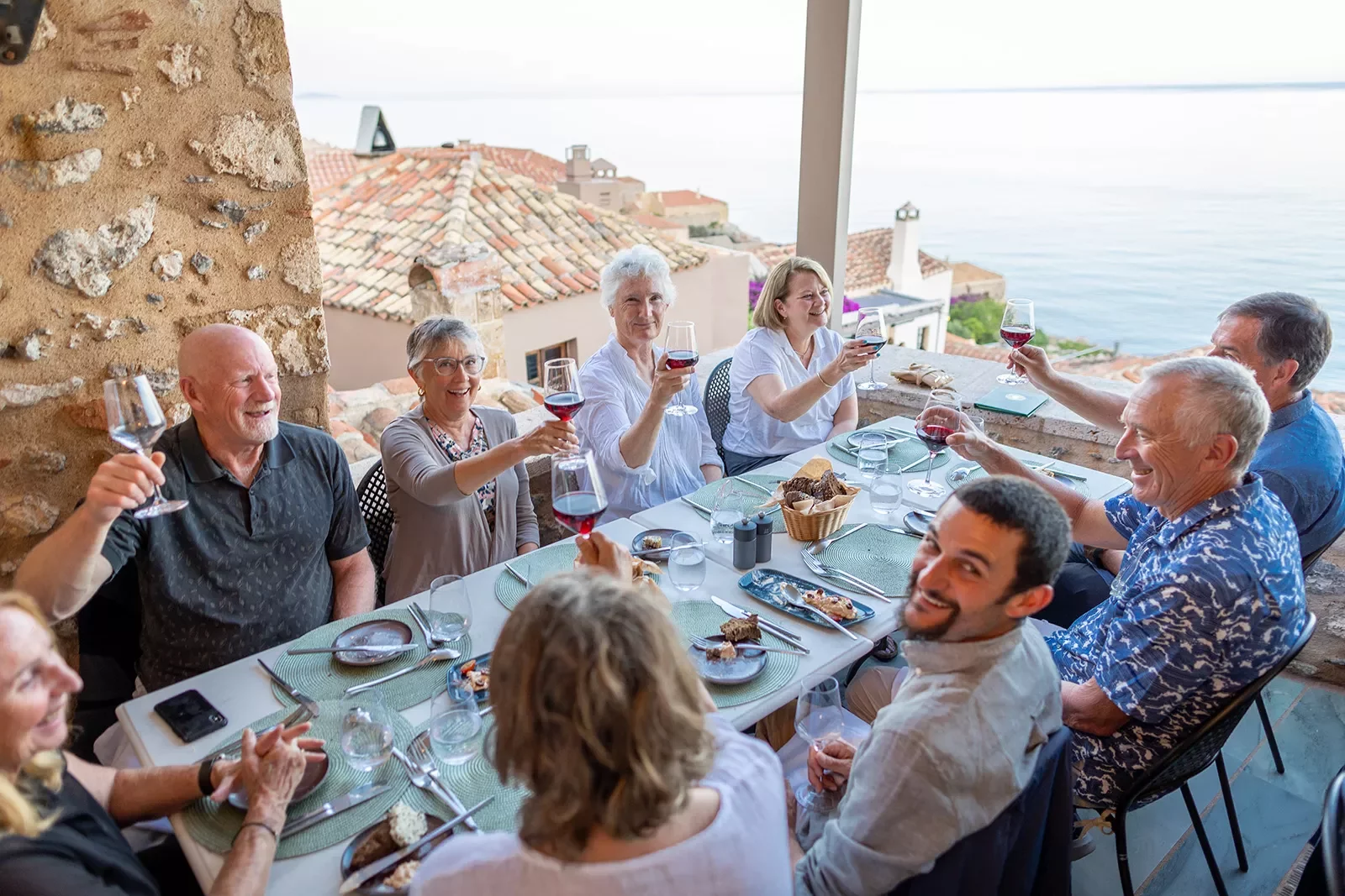 Guests at dinner table, all cheersing wine glasses, overlooking ocean.