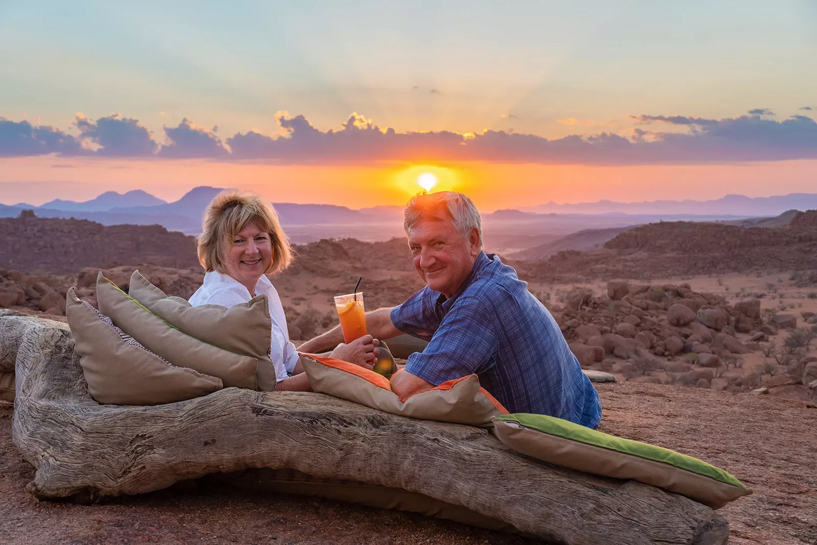A couple leaning against pillows against a fallen tree, enjoying the sunset