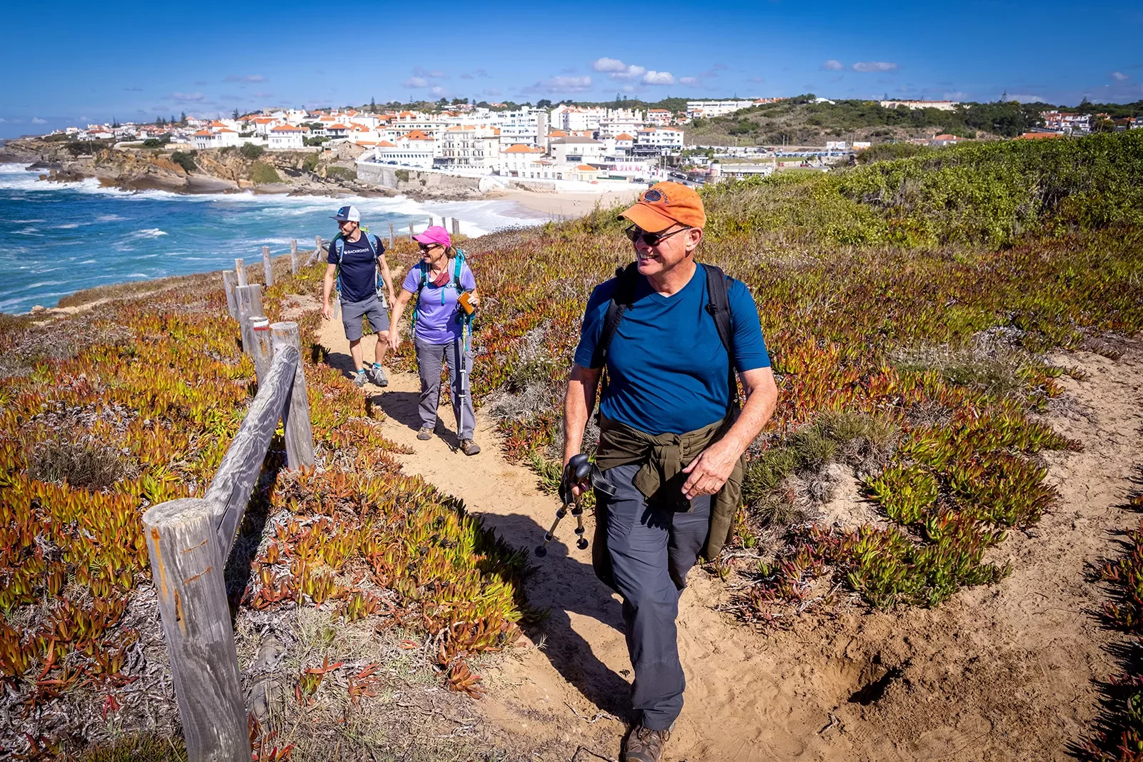 Three guests walking along ocean cliff, beach town behind them.