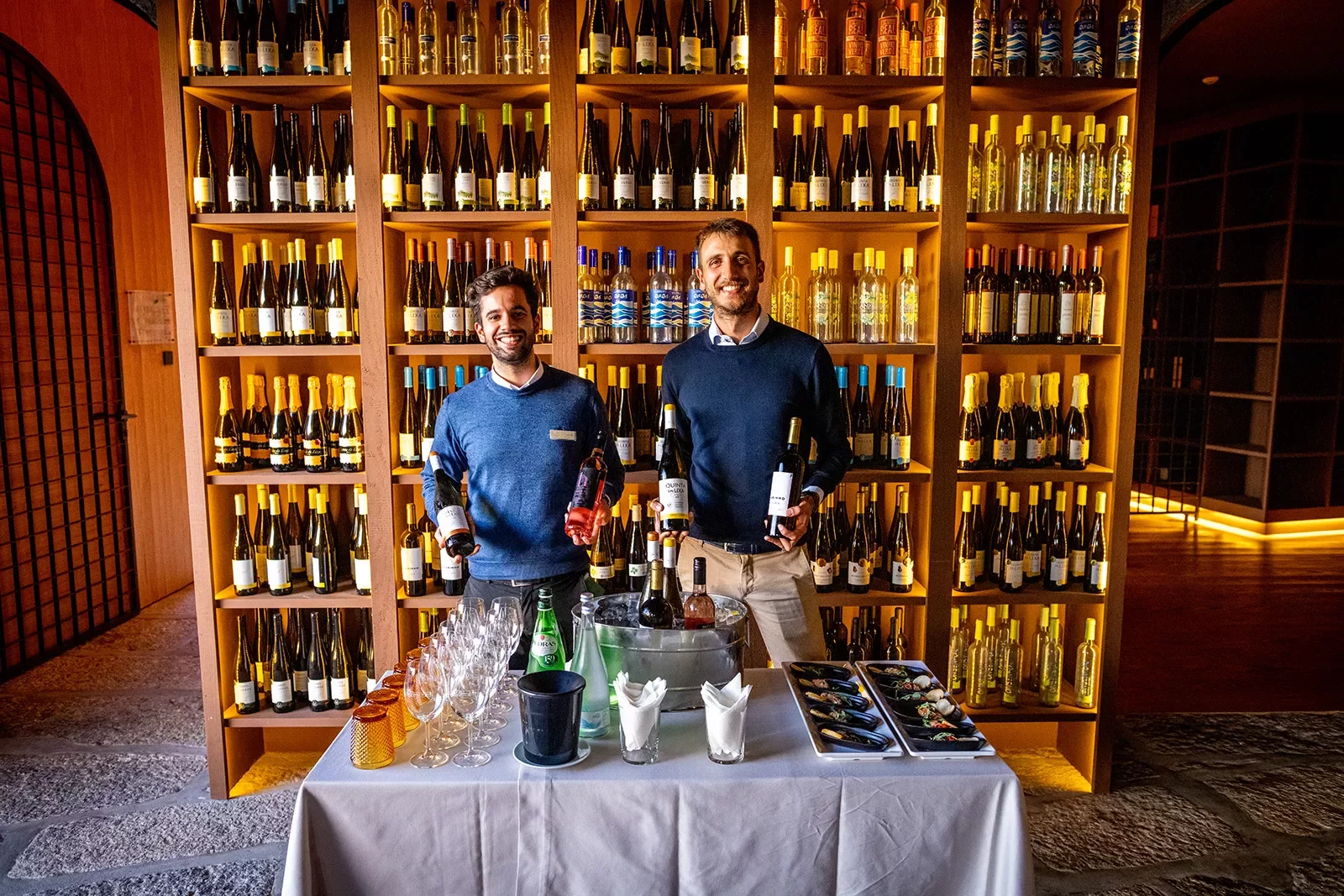 Two local wine sellers, wall of bottles behind them, table in front.