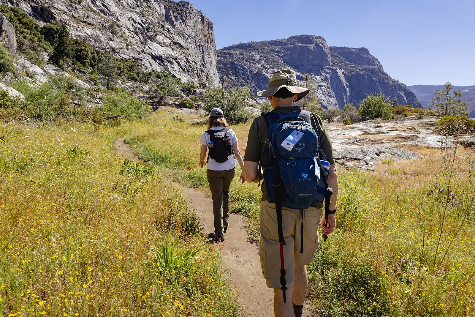 Two guests hiking on grassy trail, large cliffs to their left.