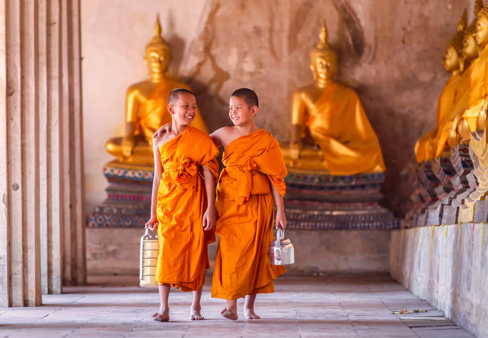 Two boys with orange monk robes, walking through a temple full of Buddhist statues