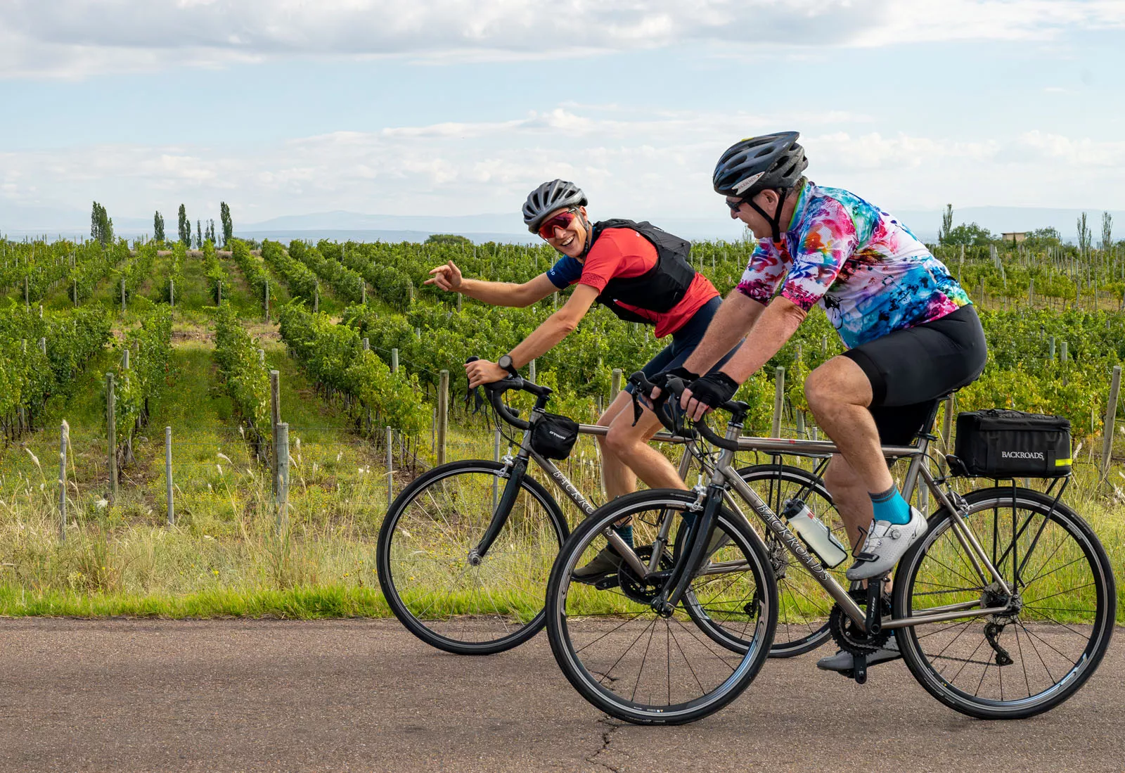Two men smiling while riding bicycles on a road along a vineyard