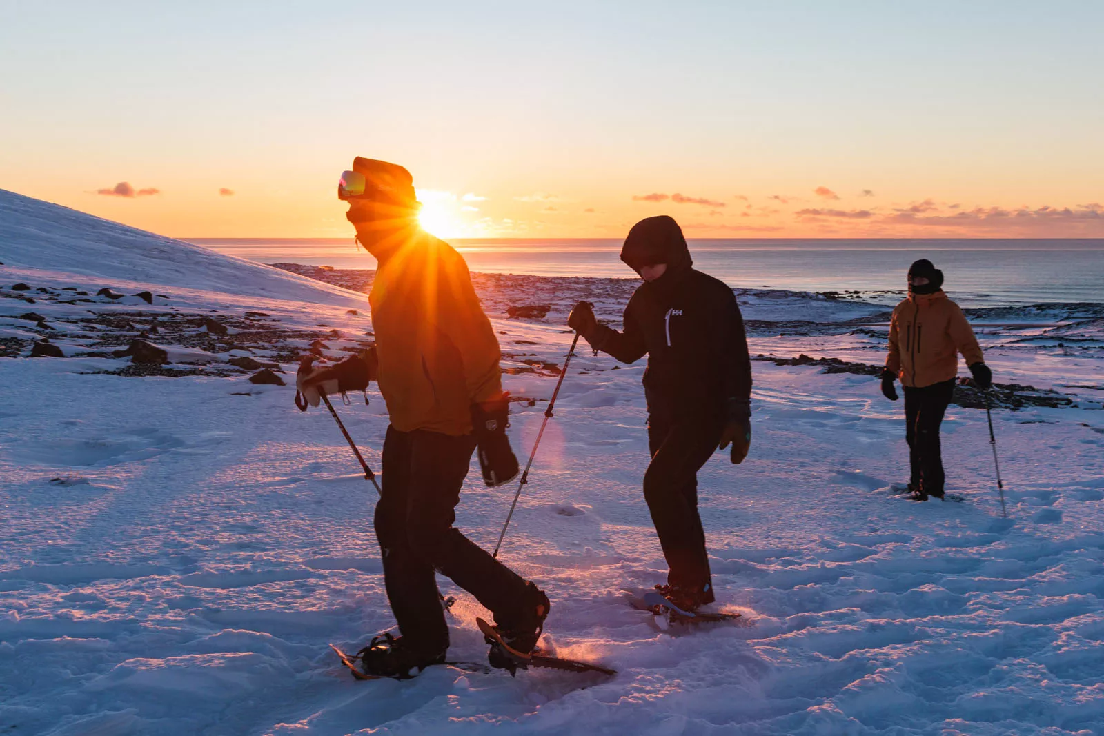 Three people wearing snow gear, walking on a field of snow by the ocean