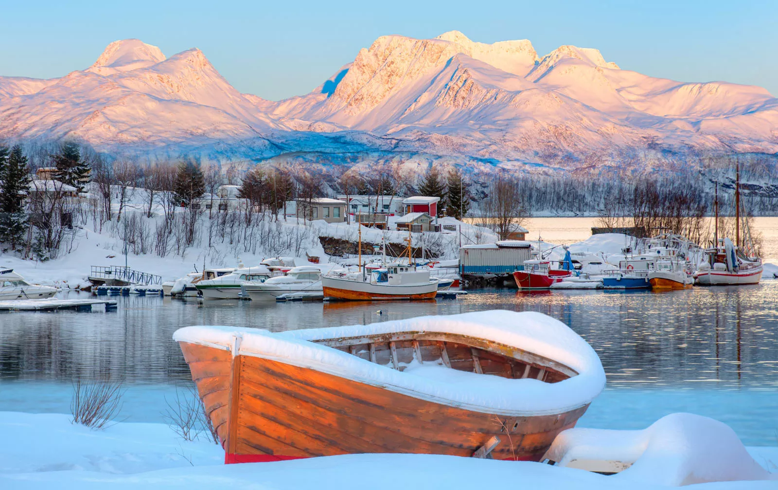 Wooden boats surrounding a lake, covered in snow with snowy mountains in the distance