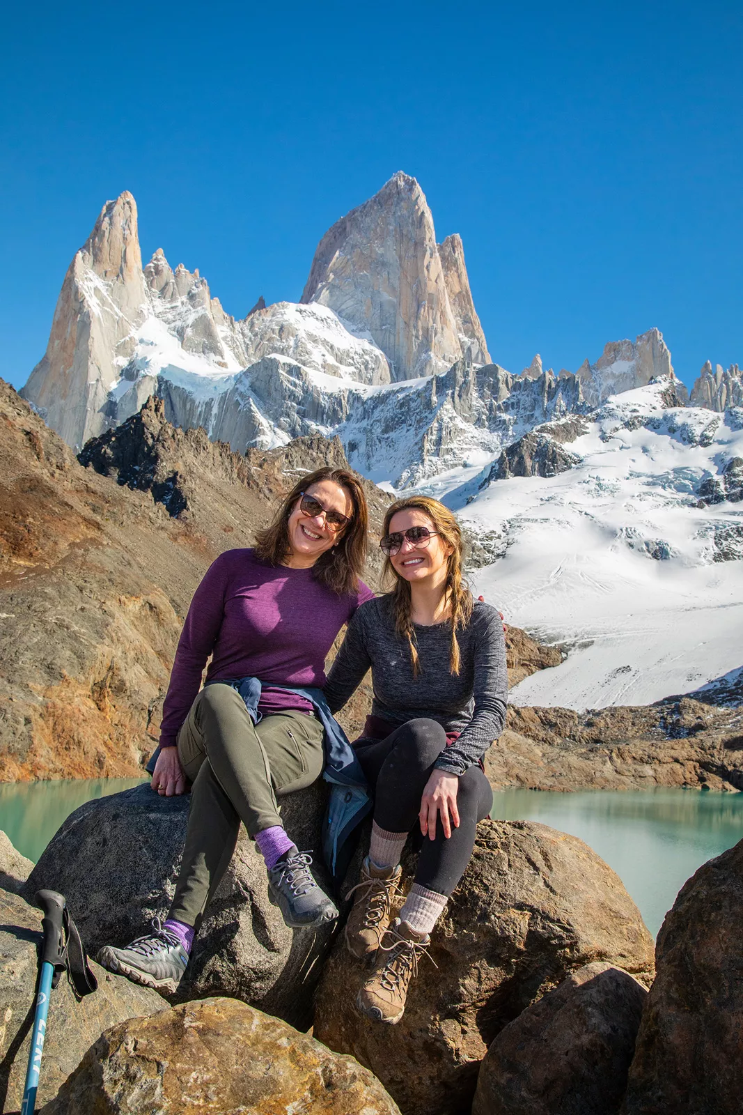 Two women smiling while sitting on boulders in front of a lake