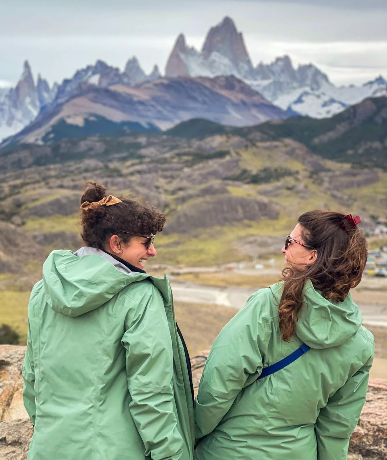 Two women wearing green jackets, smiling while looking towards an open valley