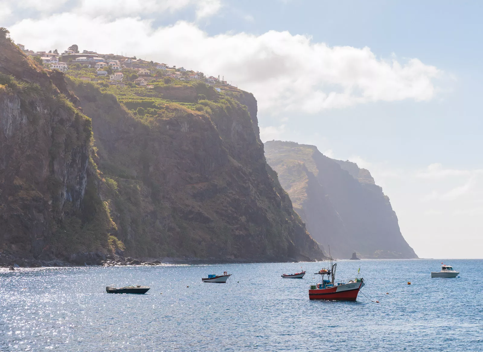 Boats floating in the ocean, with a large cliff with houses in the background