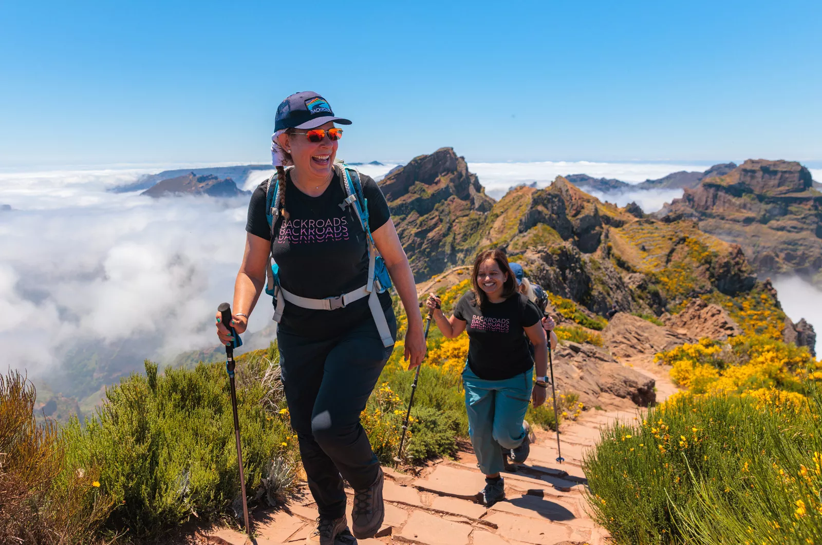 Two women smiling, while ascending a stone path on a mountain with hiking poles
