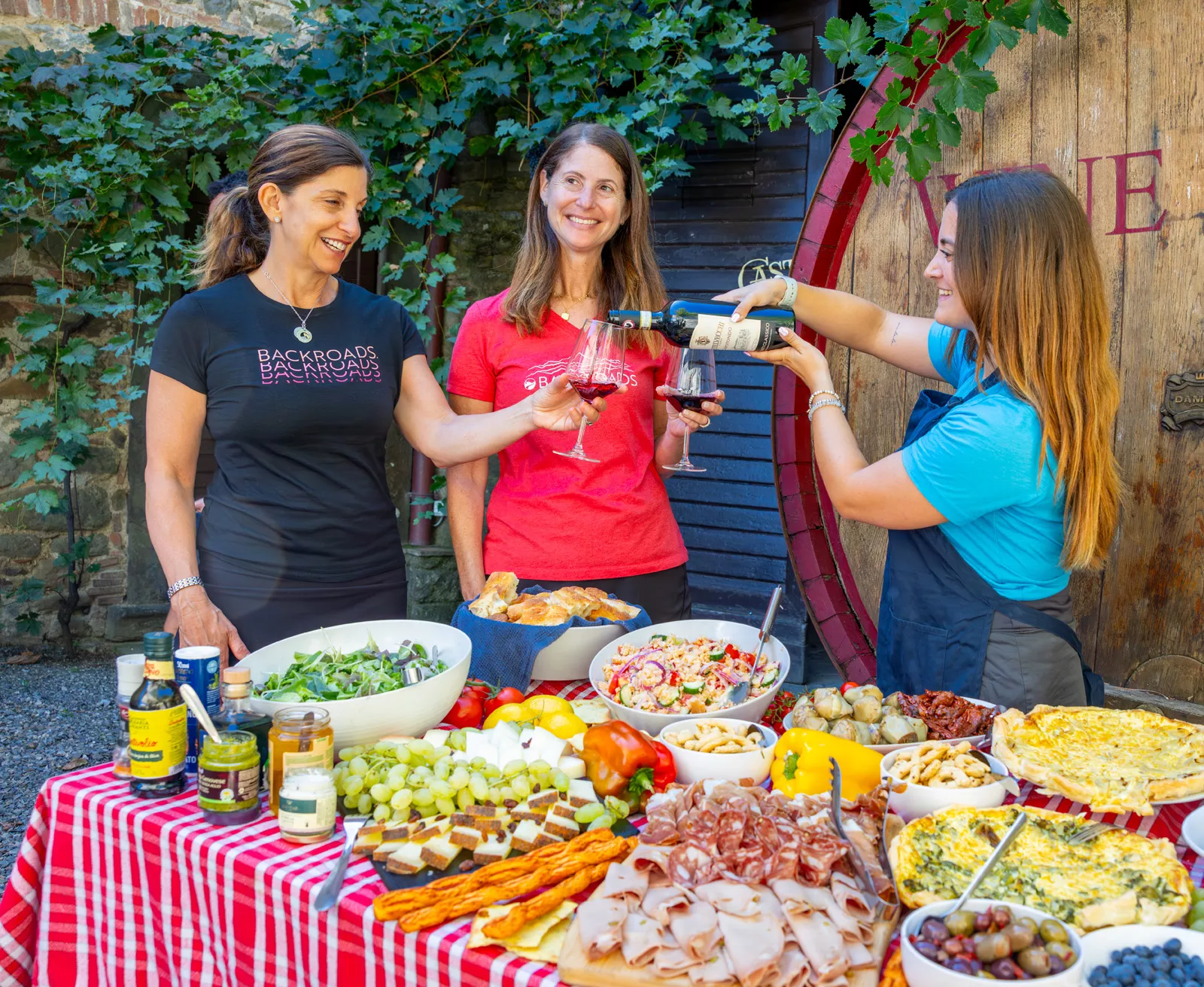 Three women behind a table full of food, pouring wine into glasses