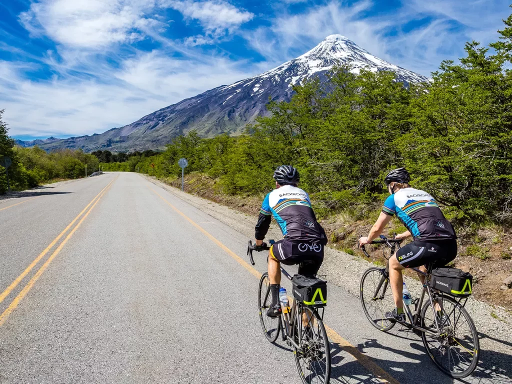 Two guests cycling down mountain road, snowy peak in background.
