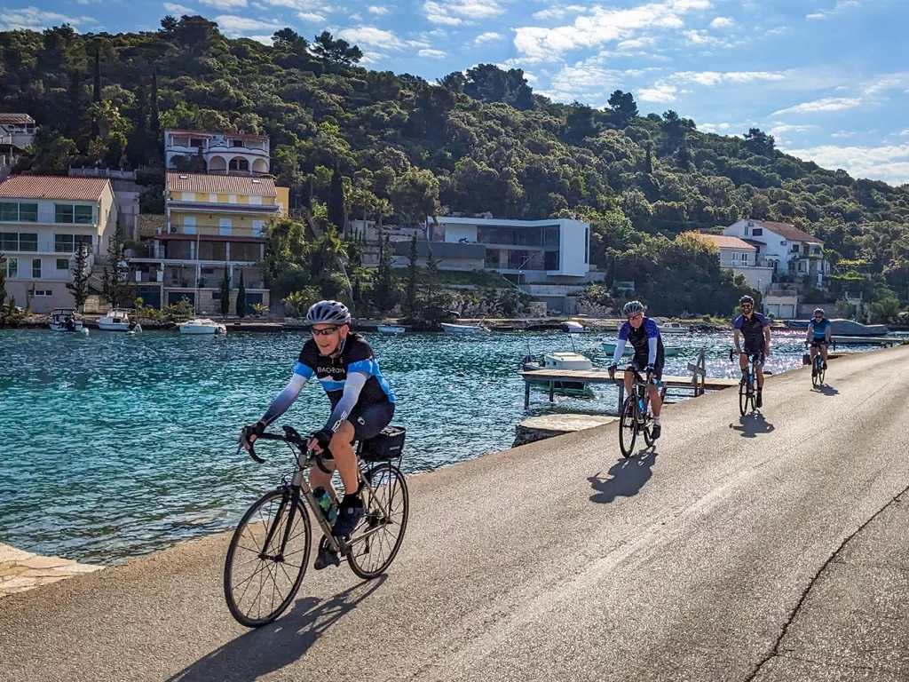 Four guests cycling down coastal road, blue ocean, hillside houses behind them.