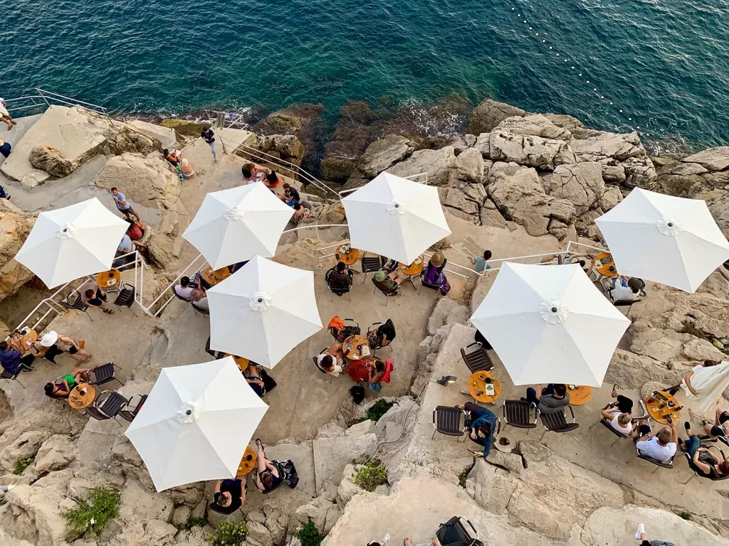Overhead shot of people eating beneath white umbrellas, stone cliffs, ocean.