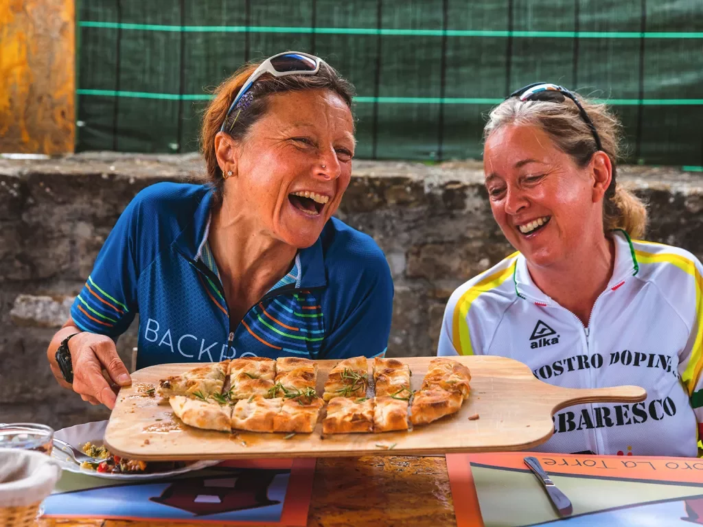 Two guests at lunch, both laughing, passing focaccia.