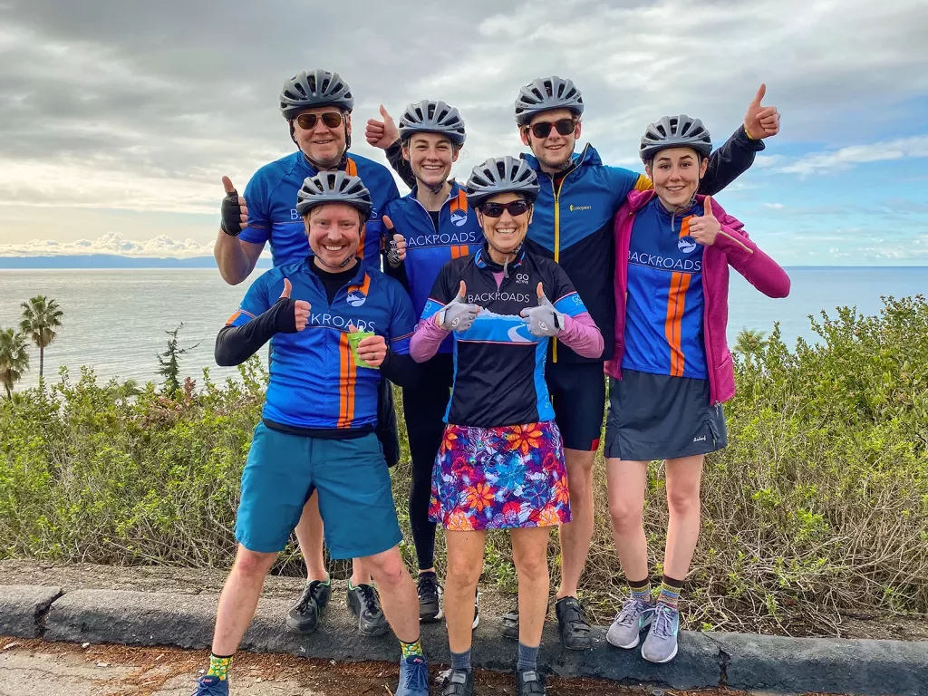 Six guests in biking gear, giving thumbs ups, ocean behind them.