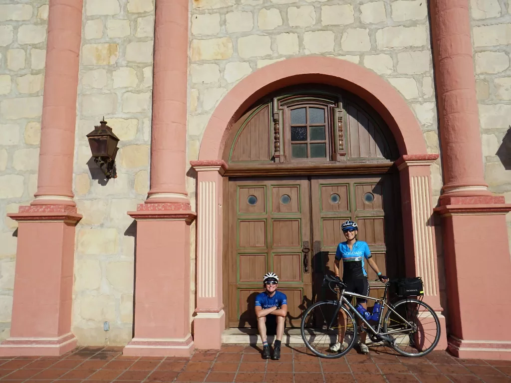 Two guests, one has their bike, in front of large wooden door. White and pink stonework around them.