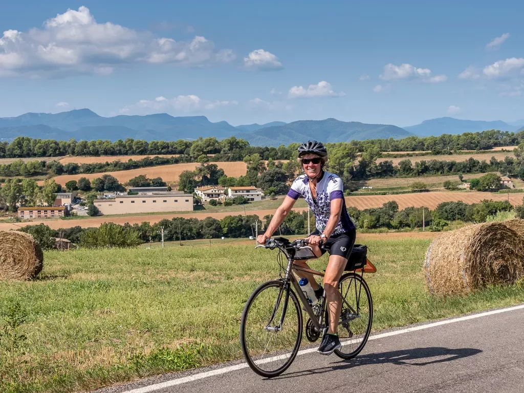 Guest cycling past farmland, hay bales, hilly vista behind her.