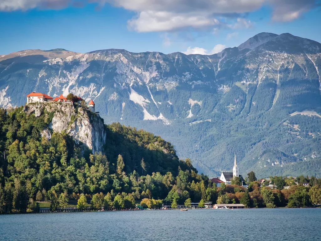 Wide shot of Bled Castle, town, lake below, mountain behind.