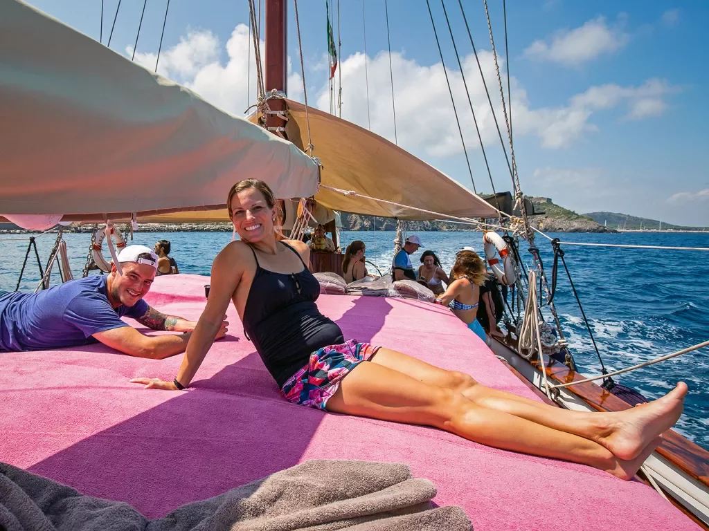 Group of guests relaxing on sailboat, coastal town in background.