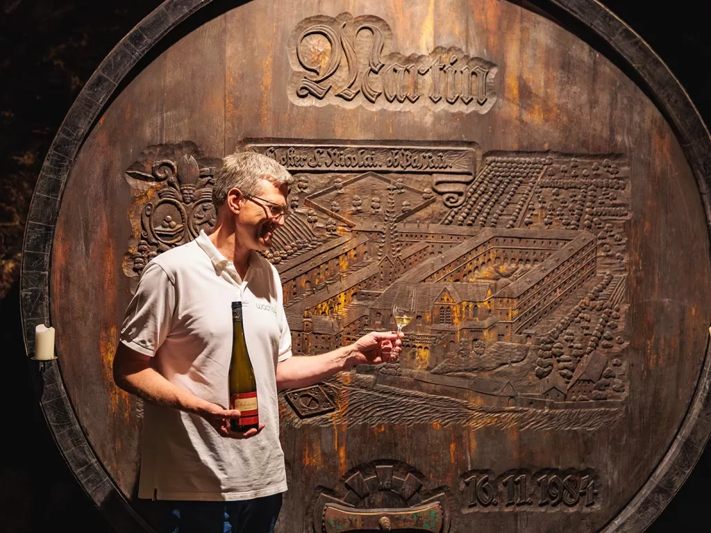 Man holding a bottle of wine in one hand and a glass with wine in the other in front of a very large wine barrel