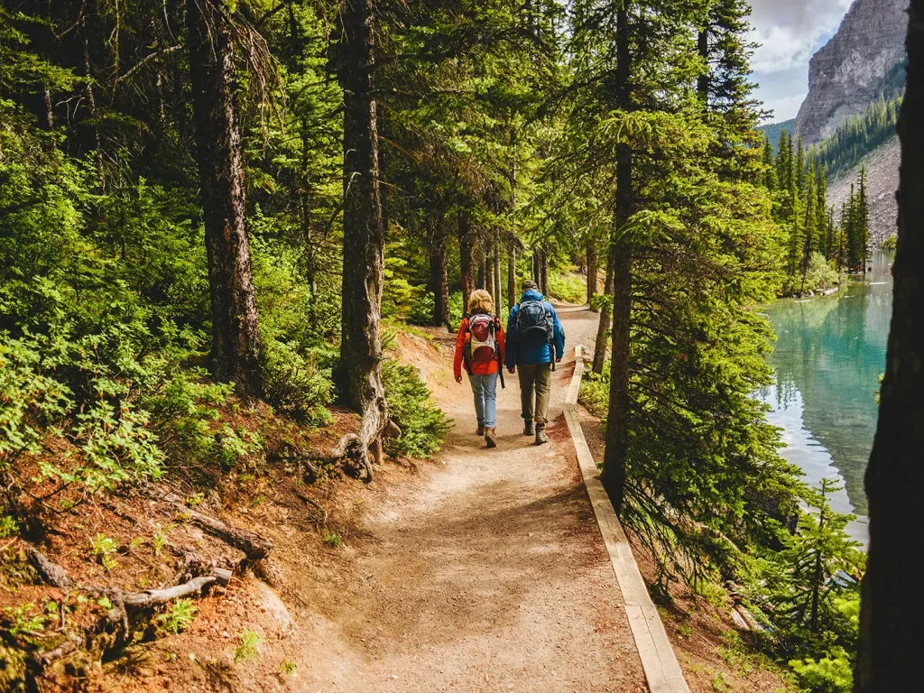 Two guests hiking down forest trail, river or lake to their right.