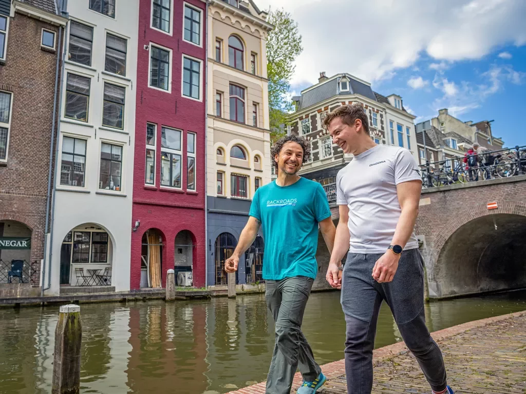 Two guests walking next to a river in town, colorful storefronts behind them.