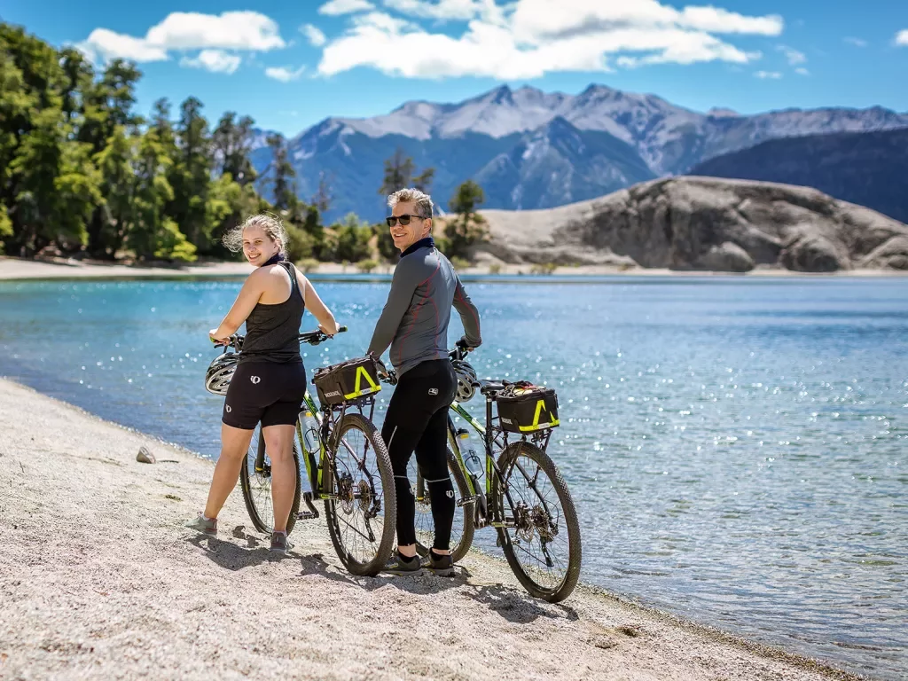 Two guests with bikes, walking along beach, blue water, forest behind them.
