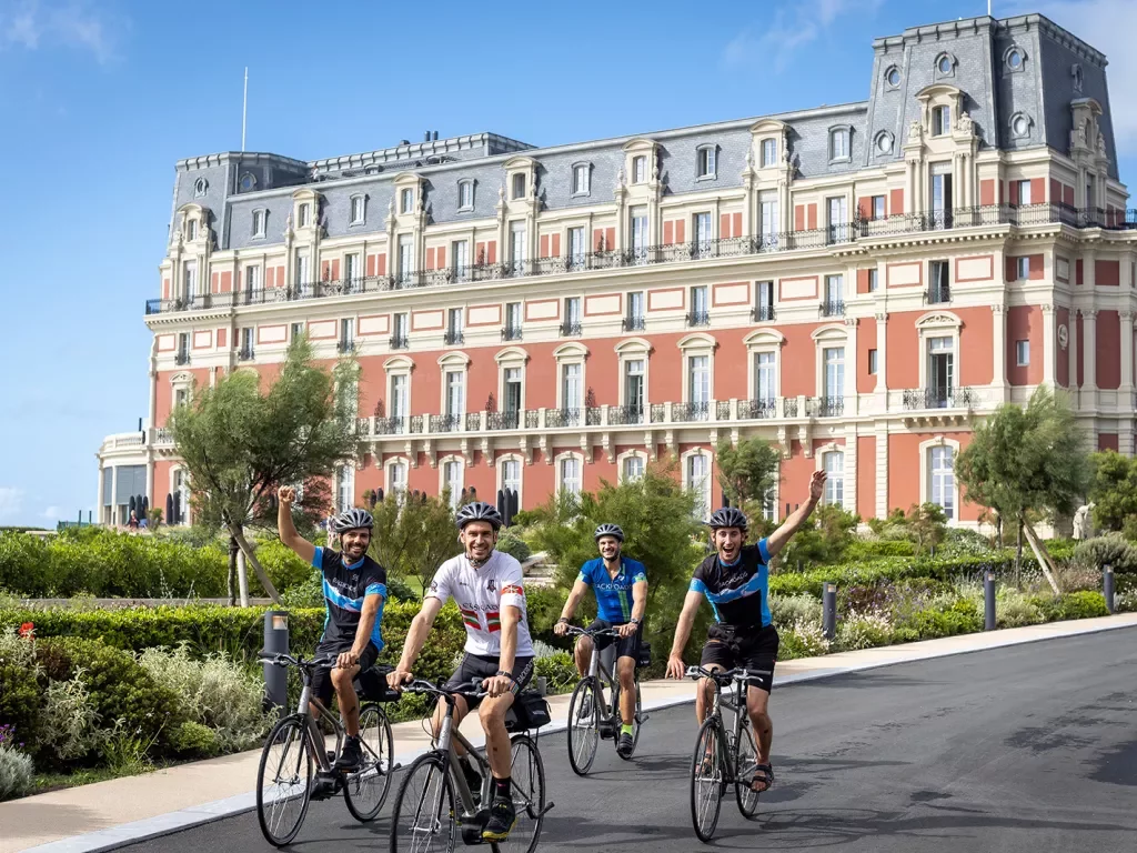 Four guests cycling past Hôtel Du Palais Biarritz, waving to camera.