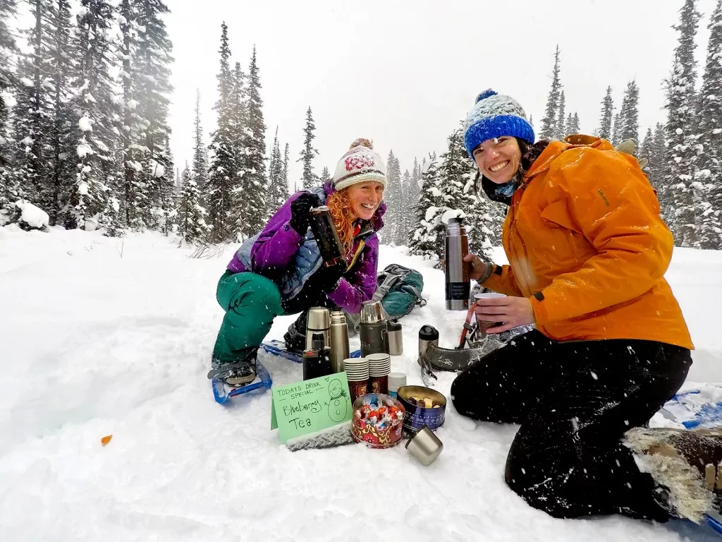 Two guests in snow, getting blueberry tea from thermos.