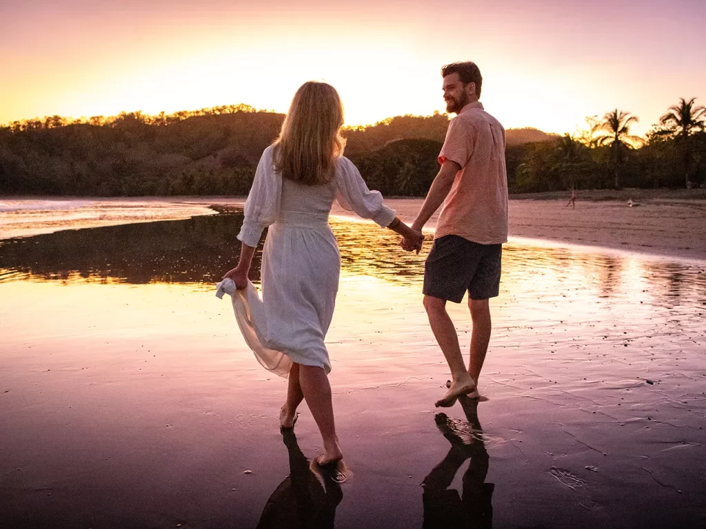 Two guests holding hands, walking along shore during sunset.