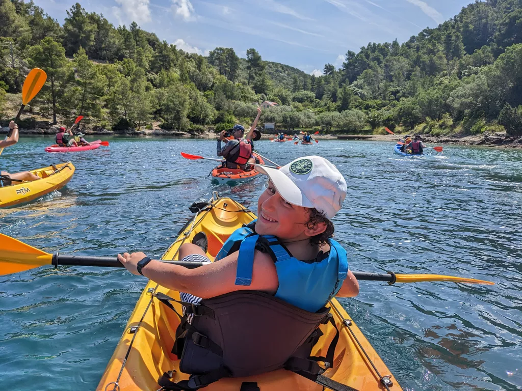 POV shot of guests kayaking, forested coast in distance.