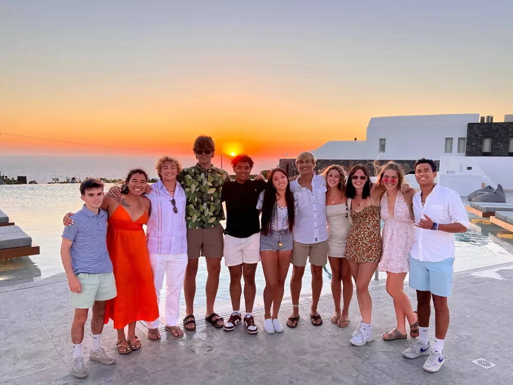 Group of young guests, arms over each other, sunset, white building behind them.