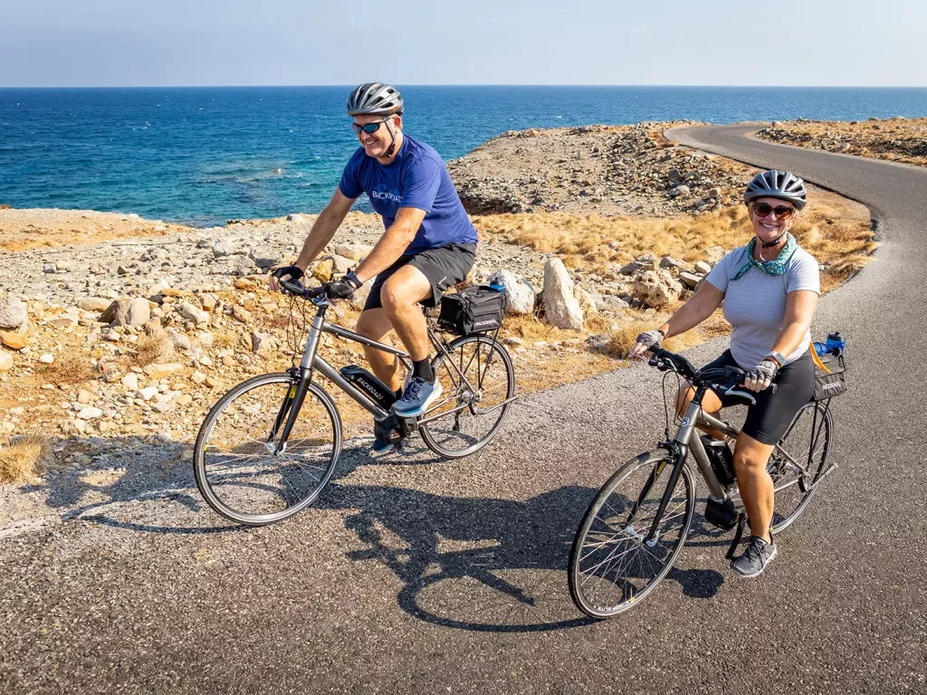 Two guests biking alongside golden coastline, smiling at camera.
