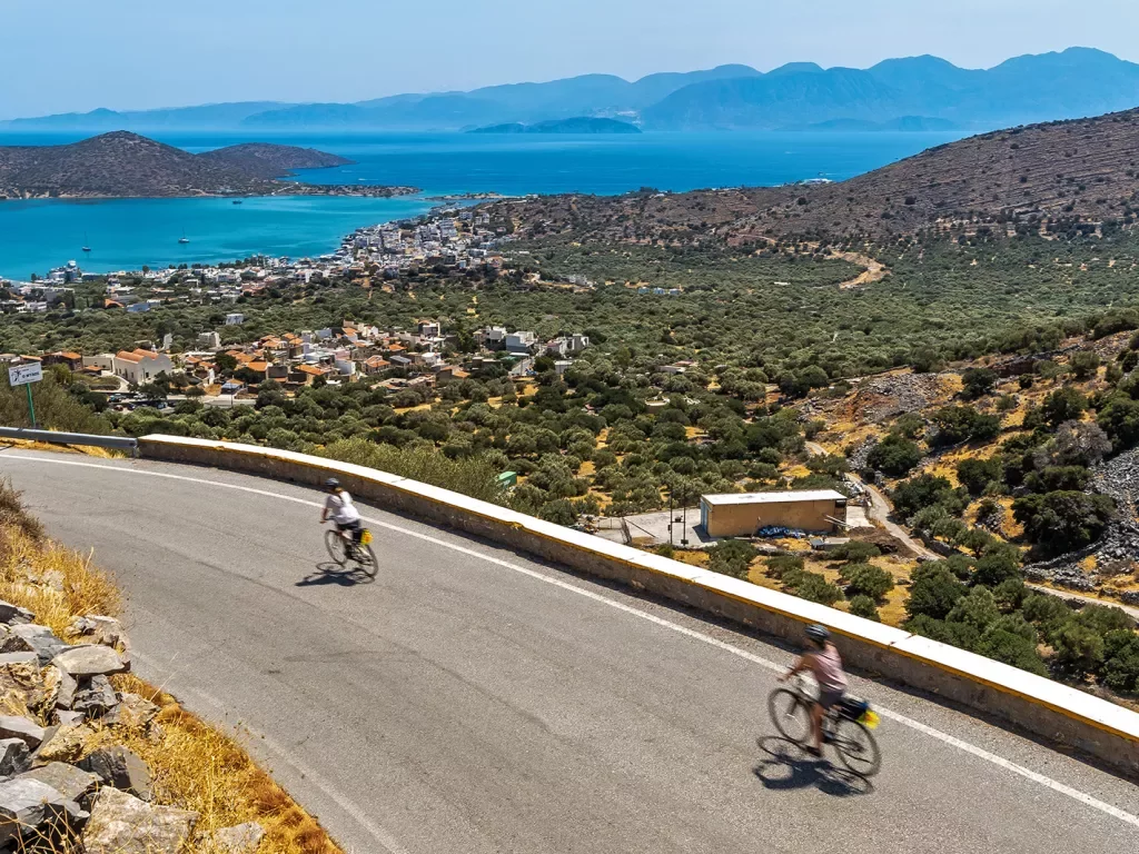 Two guests cycling down road, arid coastal town, ocean in distance. 
